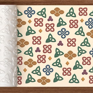 Puede incluir: Un rodillo cilíndrico blanco con un patrón en relieve de nudos celtas, triángulos y formas de diamante, mostrado junto a una superficie plana que presenta el mismo diseño en colores azul, verde, marrón y rojo sobre un fondo crema.