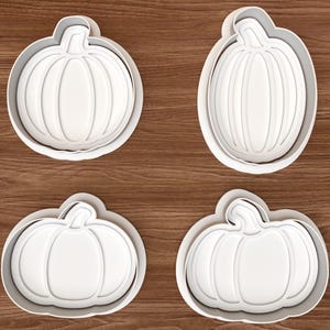 Op de afbeelding: Vier witte koekjesvormpjes in de vorm van pompoenen, gerangschikt in een raster. Elke vorm toont een uniek pompoensilhouet met een steeltje en verticale ribbels, een opstaande rand voor het snijden en een stempelontwerp aan de binnenzijde.