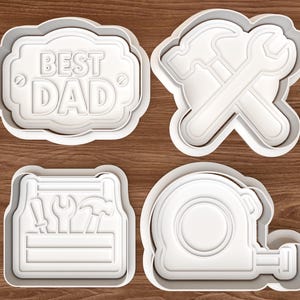 May include: Four white plastic cookie cutters. One says "BEST DAD". The others are shaped like a hammer and wrench, a toolbox with tools, and a tape measure. The cutters are on a wooden surface.