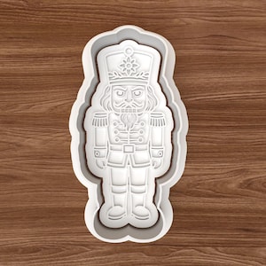 Könnte beinhalten: Ein weißer Ausstecher aus Kunststoff in Form eines Nussknacker-Soldaten. Der Ausstecher hat detaillierte Linien für Uniform, Gesicht und Hut. Der Ausstecher befindet sich auf einer Holzoberfläche.