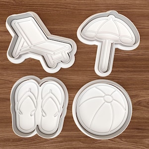 Könnte beinhalten: Vier weiße Ausstecher aus Kunststoff in Form von Sommerartikeln. Die Formen umfassen einen Strandstuhl, einen Sonnenschirm, ein Paar Flip-Flops und einen Strandball. Die Ausstecher liegen auf einer braunen Holzoberfläche.