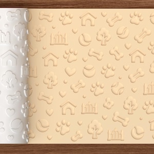 May include: A white cylindrical rolling pin with an embossed pattern of dog-related icons including paw prints, bones, tennis balls, trees, hearts, and dog houses, shown alongside a light tan sheet of dough featuring the same imprinted design.