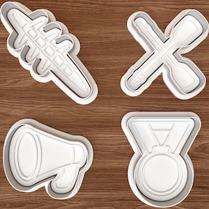 Pode incluir: Um conjunto de quatro cortadores de biscoitos de plástico branco com formas distintas: um objeto longo segmentado, dois remos cruzados, um megafone e uma medalha circular com fita. Cada cortador tem uma borda elevada para cortar a massa.
