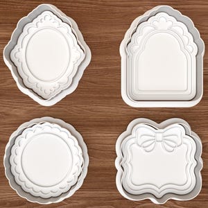 Puede incluir: Un juego de cuatro cortadores de galletas de plástico blanco con formas variadas, incluyendo un óvalo con borde festoneado, un marco arqueado, un diseño circular festoneado y un marco rectangular con un lazo, todos con relieves.