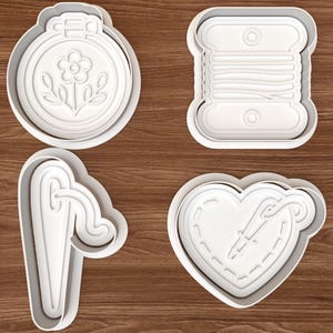 Può includere: Un set di quattro tagliabiscotti in plastica bianca con motivi legati al cucito: un telaio da ricamo circolare con un fiore, una bobina di filo rettangolare, un lungo ago da cucito con filo e un cuore con ago e filo.