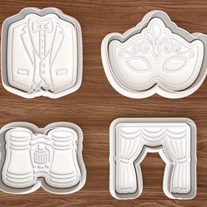 May include: Four white cookie cutters in the shapes of a tuxedo jacket, a masquerade mask, opera glasses, and theater curtains. The cutters are on a wooden surface. These cutters are ideal for baking themed cookies.