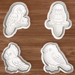 Könnte beinhalten: Vier weiße Ausstecher aus Kunststoff in Form von Vögeln. Das Set enthält einen Papagei auf einem Ast, einen Wellensittich auf einer Stange, einen Kakadu und einen kleinen Vogel. Die Ausstecher liegen auf einer braunen Holzoberfläche.