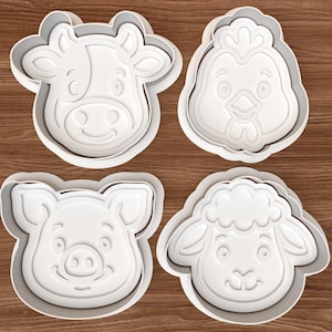 May include: Four white plastic cookie cutters in the shapes of farm animals: a cow, a chicken, a pig, and a sheep. The cutters have a detailed design of each animal's face and are set against a wooden background.