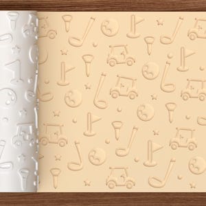 May include: A white cylindrical rolling pin with a repeating embossed pattern of golf carts, golf clubs, golf balls, tees, flags, and stars, shown next to a light tan dough sheet featuring the same golf-themed design pressed into its surface.