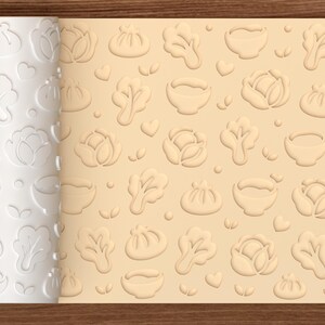Könnte beinhalten: Eine weiße zylindrische Teigrolle mit einem wiederkehrenden geprägten Muster aus Teigtaschen, Schüsseln und Blattgemüse, die das gleiche Design in eine flache, hellbeige Teigoberfläche drückt. Ein kleines Keks-Symbol ist in der Ecke zu sehen.