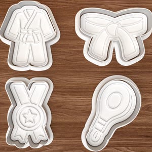 May include: Four white plastic cookie cutters on a wooden surface. The cutters are shaped like a karate uniform, a bow, a medal with a star, and a tennis racket. The cutters are designed for making biscuits.