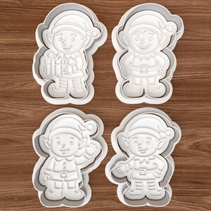 May include: Four white plastic cookie cutters in the shape of cartoon elves. Each elf has a hat, belt, and shoes. One elf holds a gift, another waves, and the others have open hands. The cutters are on a wooden surface.