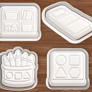 May include: Four white plastic cookie cutters featuring school-themed designs. One cutter shows a shelf with bins and blocks, another a rectangular pencil case, one a cup filled with crayons, and the last a board with circle, square, triangle, and hexagon shapes.
