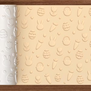 Peut inclure: Un rouleau à pâtisserie cylindrique blanc présentant un motif en relief répétitif composé d'œufs de Pâques, de lapins, de carottes, de tulipes et de paniers. Le rouleau est placé à côté d'une surface beige claire portant le même motif.