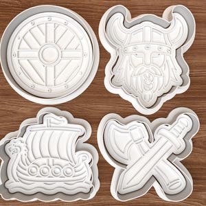 May include: Four white plastic cookie cutters in Viking theme. The shapes include a shield, a Viking helmet, a longboat, and crossed axe and sword. The cookie cutters are on a wooden surface.