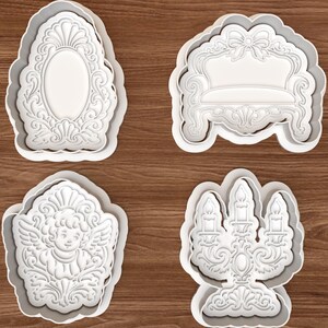 May include: A set of four white plastic cookie cutters featuring intricate embossed designs: an ornate oval mirror, a detailed sofa with a bow, a cherub with wings, and a three-armed candelabra with lit candles, all with scalloped outer edges.