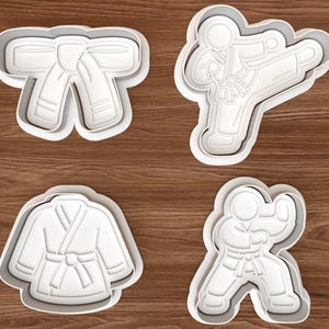 May include: Four white plastic cookie cutters in karate-themed shapes. The designs include a karate belt, a karate uniform, and two karate figures in action poses. The cutters are on a brown wooden surface.