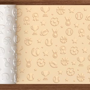 May include: A cylindrical rolling pin with a white surface featuring an embossed pattern of sports-themed icons including trophies, medals, stars, basketballs, baseballs, and footballs, shown alongside a flat beige dough imprint of the same design.