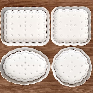 Puede incluir: Cuatro cortadores de galletas de plástico blanco en diferentes formas, incluyendo cuadrados y ovalados, con bordes festoneados y detalles de puntos. Dispuestos sobre una superficie de madera, para hornear.