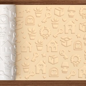 May include: A white cylindrical rolling pin with a repeating embossed pattern of houses with heart windows, potted plants, keys, stars, lamps, wreaths, and boxes, shown next to a light tan dough sheet displaying the same imprinted design.