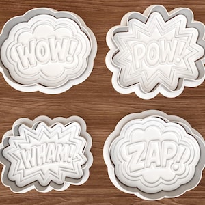 May include: A set of four white plastic cookie cutters in comic book speech bubble shapes. The cutters feature the embossed words WOW!, POW!, WHAM!, and ZAP! with distinct cloud-like and jagged outlines for each individual design element.