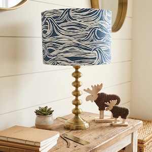 Op de afbeelding: Een tafellamp met een messingkleurige voet van gestapelde bollen en een cilindrische lampenkap met een donkerblauw en wit grafisch patroon van gestileerde oceaangolven. De lamp staat op een houten ondergrond.