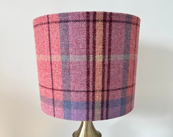 May include: A lampshade with a plaid pattern in shades of pink, purple, and blue. The lampshade is cylindrical and sits atop a gold-colored lamp base. The plaid design features vertical and horizontal lines.
