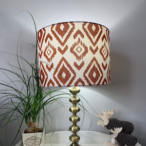 Puede incluir: Una lámpara de mesa dorada con una pantalla de lámpara de patrón geométrico blanco y naranja. La lámpara está sobre una mesa blanca con una planta en maceta y dos figuritas de alce de madera.