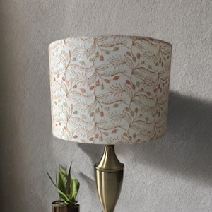 Könnte beinhalten: Eine dekorative Tischlampe mit einem weißen Lampenschirm mit einem sich wiederholenden Muster aus braunen und grünen Blättern und Beeren. Die Lampe hat einen messingfarbenen Sockel und einen zylindrischen Schirm. Eine kleine Topfpflanze steht neben der Lampe.