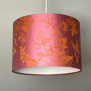 Puede incluir: Pantalla de lámpara cilíndrica con un estampado floral. La pantalla tiene un degradado de color de rosa a burdeos con diseños florales naranjas. La pantalla cuelga de un cable blanco y tiene un interior blanco.