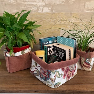 Puede incluir: Tres cestas de almacenamiento de tela con patrones florales. Dos cestas están llenas de plantas, y la tercera cesta contiene libros y un mando a distancia.