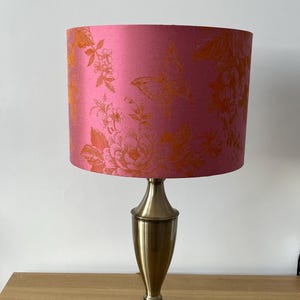 May include: A table lamp with a pink lampshade featuring an orange floral and bird pattern. The lamp has a brass-colored base. The lampshade is cylindrical and the base has a classic, elegant design.