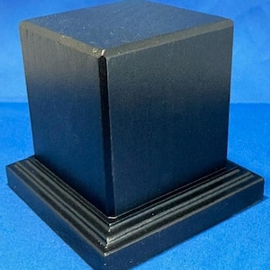 May include: A black wooden pedestal with a square top and a base with three layers of moulding.