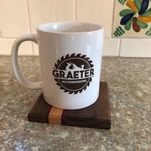 May include: White ceramic coffee mug with a brown logo that says "Graeter Woodworking" inside a circular saw blade design. The mug is sitting on a wooden coaster with a walnut and maple wood design.