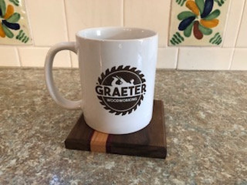 May include: White ceramic coffee mug with a brown logo that says "Graeter Woodworking" inside a circular saw blade design. The mug is sitting on a wooden coaster with a walnut and maple wood design.