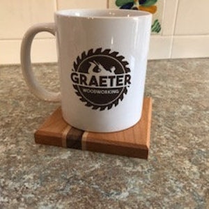 May include: White ceramic mug with a brown logo that says "Graeter Woodworking" inside a circular saw blade design. The mug is sitting on a wooden coaster with a thin stripe of light wood.