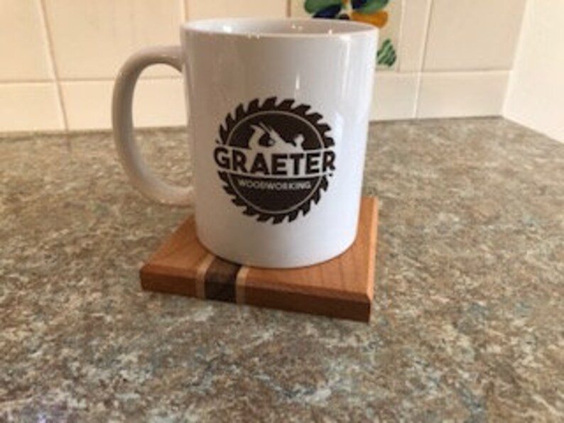 May include: White ceramic mug with a brown logo that says "Graeter Woodworking" inside a circular saw blade design. The mug is sitting on a wooden coaster with a thin stripe of light wood.