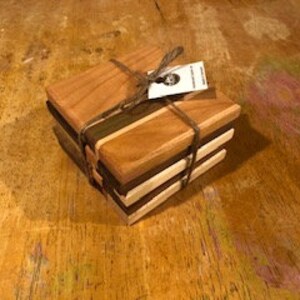 May include: Set of four wooden coasters, made from cherry, walnut, and maple wood. The coasters are square and have a natural wood finish. They are stacked on top of each other and tied together with twine.