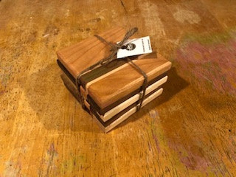 May include: Set of four wooden coasters, made from cherry, walnut, and maple wood. The coasters are square and have a natural wood finish. They are stacked on top of each other and tied together with twine.