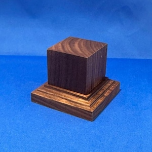 May include: A small, square wooden pedestal with a dark brown finish. The pedestal has a tiered base with three layers.