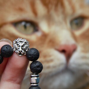 Peut inclure: Un bracelet en pierre de lave noire avec un pendentif tête de lion argenté. Le bracelet est tenu par une main, avec un chat orange flou en arrière-plan.