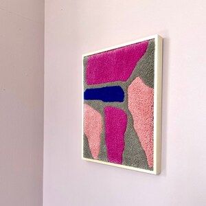 May include: A framed abstract wall art piece with a textured, fuzzy surface. The artwork features a variety of shapes in shades of pink, gray, and blue.