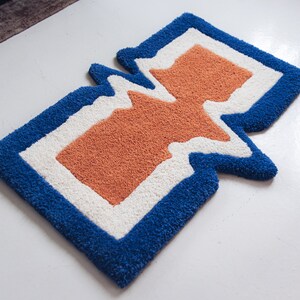 Puede incluir: Una alfombra azul, blanca y naranja con un dise&ntilde;o abstracto &uacute;nico. La alfombra tiene una forma ondulada, org&aacute;nica y est&aacute; hecha de un material suave y mullido.