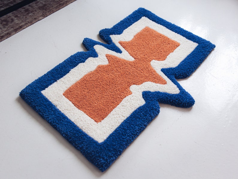 Puede incluir: Una alfombra azul, blanca y naranja con un dise&ntilde;o abstracto &uacute;nico. La alfombra tiene una forma ondulada, org&aacute;nica y est&aacute; hecha de un material suave y mullido.