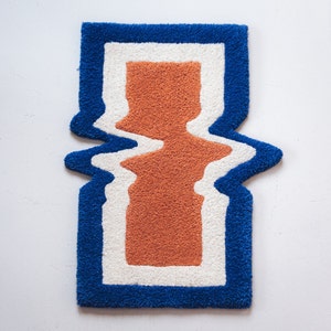 Puede incluir: Una alfombra azul, blanca y naranja con un dise&ntilde;o abstracto ondulado. La alfombra tiene una pila gruesa y texturizada.