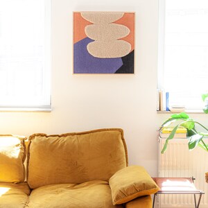 May include: A yellow velvet sofa with a throw pillow in front of a white wall with a framed abstract art piece featuring a beige, orange, and purple color scheme.