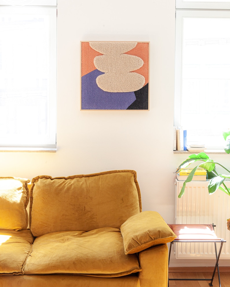 May include: A yellow velvet sofa with a throw pillow in front of a white wall with a framed abstract art piece featuring a beige, orange, and purple color scheme.