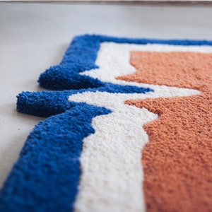 Puede incluir: Primer plano de una alfombra azul, blanca y naranja con un patr&oacute;n geom&eacute;trico. La alfombra est&aacute; hecha de un material suave y esponjoso.