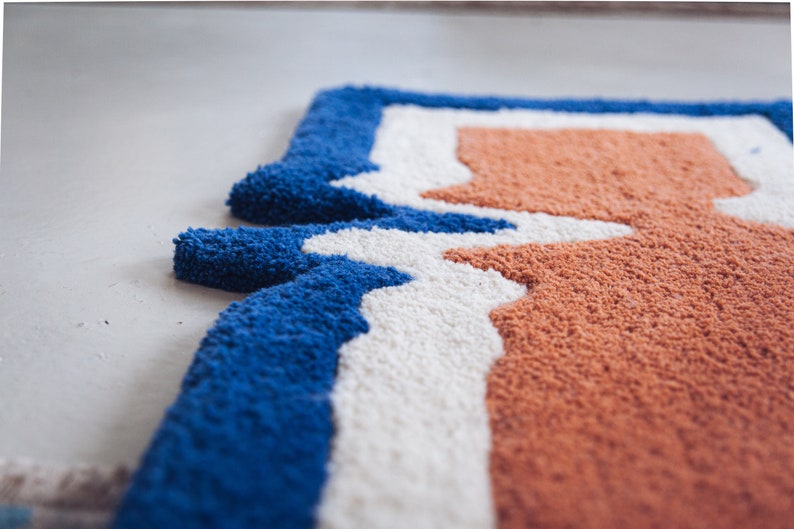 Puede incluir: Primer plano de una alfombra azul, blanca y naranja con un patr&oacute;n geom&eacute;trico. La alfombra est&aacute; hecha de un material suave y esponjoso.