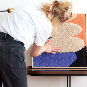 May include: A framed abstract wall art piece with a textured, multi-colored design. The artwork features a beige, orange, and purple color scheme with a black outline.